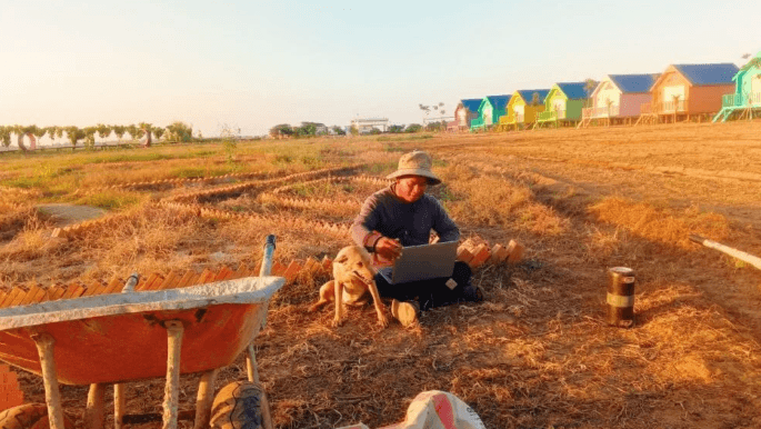 A man wearing a hat sits on dry grass working on a laptop. A light brown dog sits beside him, looking forward. In the background are colorful, small riverside houses under a bright, golden sunset sky.