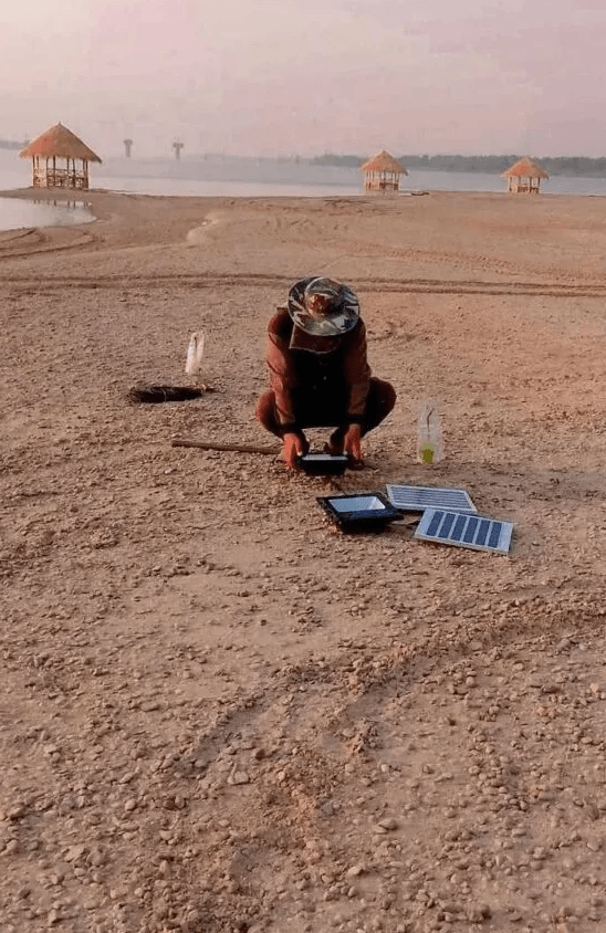 A person crouching on a wide, dry sandy beach, focused on small electronic solar panels resting on the sand. In the far distance, several small thatched-roof huts (gazebos) are visible by the water's edge under a hazy sky