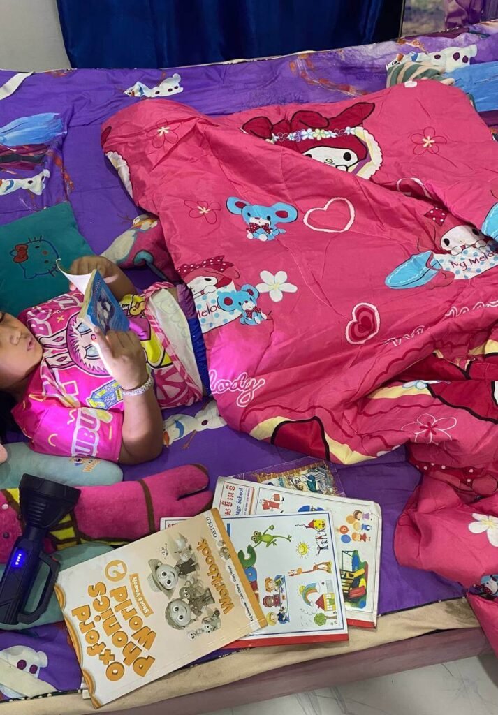 A young girl reading a phonics book in bed using a flashlight under a pink blanket.