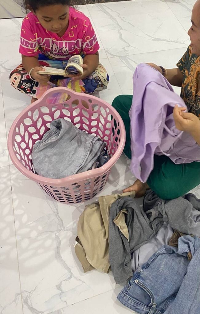 A mother folding clothes from a laundry basket while her young daughter sits on the floor reading a book aloud to her.