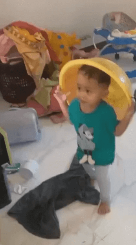 A 3-year-old boy wearing a yellow plastic colander on his head like a helmet in a messy room.